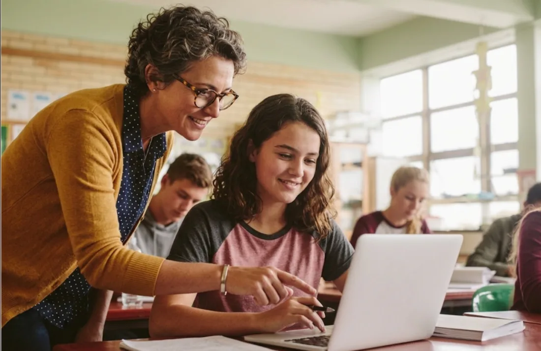 A teacher helping a student review work on a laptop in a classroom